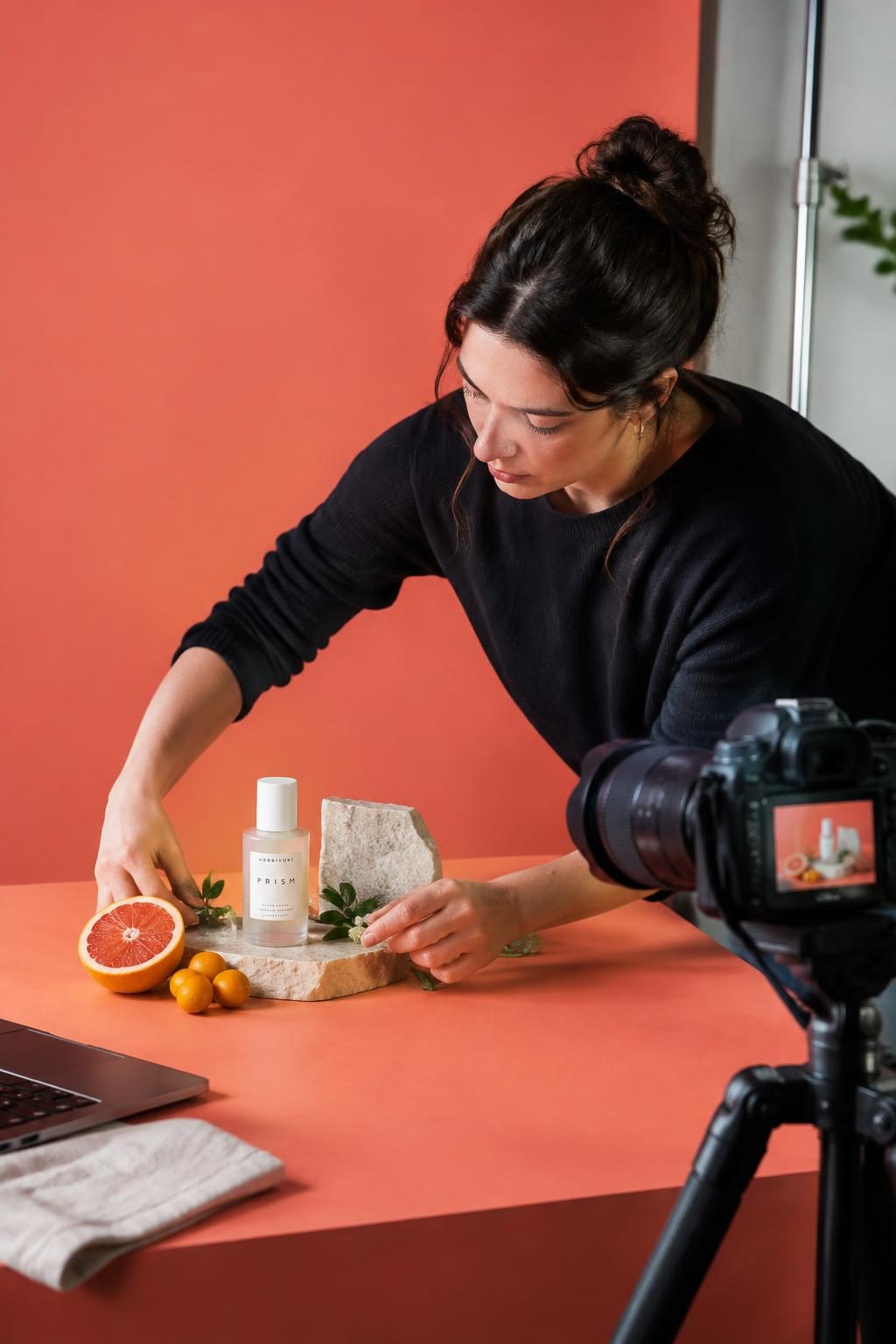 A product photographer arranges lipstick and sculptural props against an orange studio backdrop.